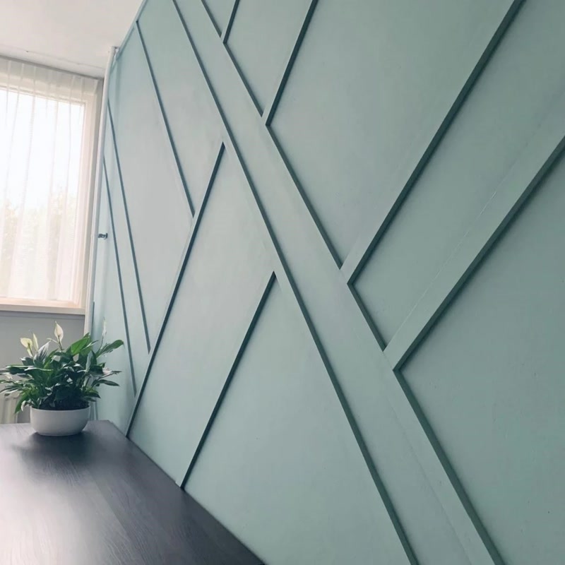 Modern adaçayı yeşili duvarda asimetrik ve geometrik desenli poliüretan çıta uygulaması ve masa üzerindeki bitki.