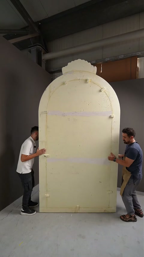 Two craftsmen carry unfinished arch backing across the production workshop.