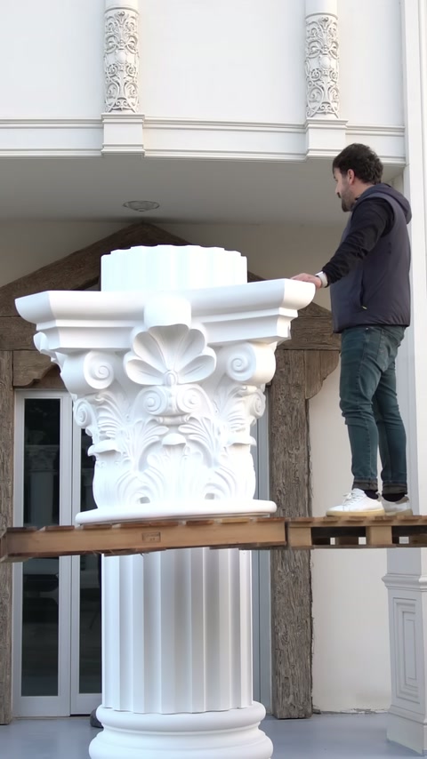 A worker on the scaffolding is fitting the upper section of a Corinthian capital onto the column.