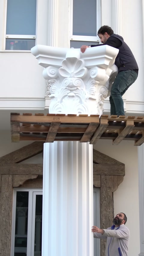 Two workers are adjusting the ornate column capital together along the shaft axis.