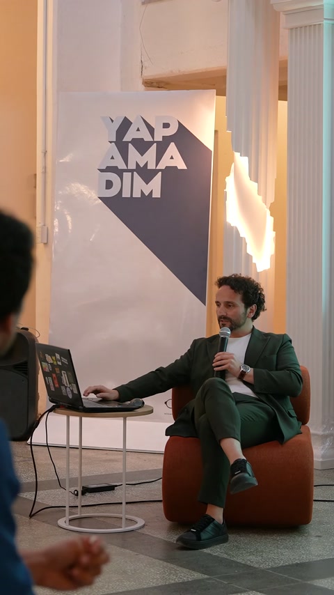 Konuşmacı, etkinlik afişi önünde oturarak dizüstü bilgisayardan sunum yapıyor