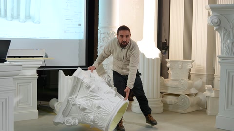 Il presentatore esamina da vicino un grande capitello di colonna con decorazioni fogliari.