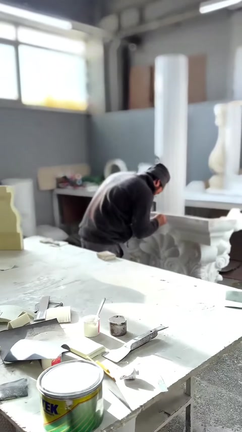Un artigiano lavora la superficie di un capitello corinzio grigio sul banco da lavoro.