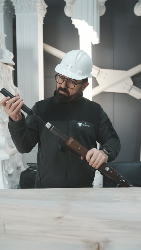 Uygulayıcı beyaz sütunların önünde tüfeğin namlusuna bakıyor.
