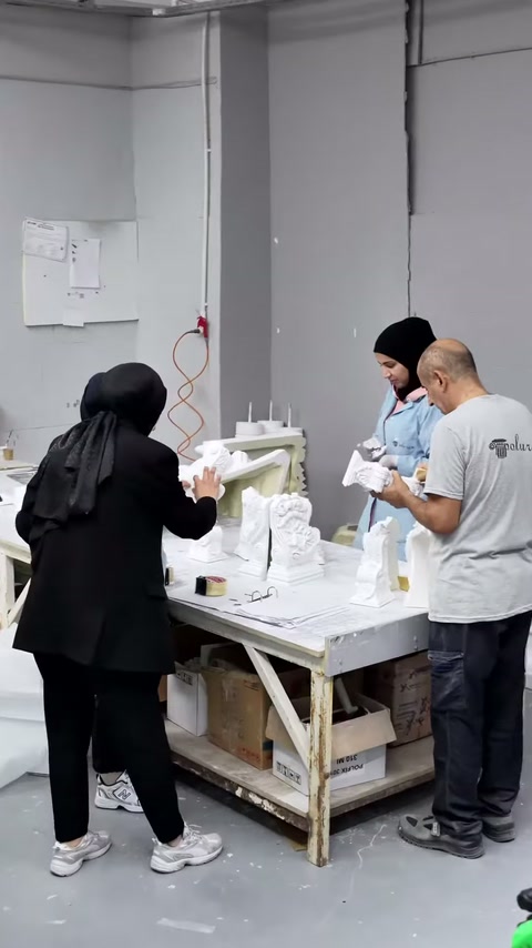 Il presentatore esamina il pezzo bianco al banco mentre i dipendenti continuano la produzione.