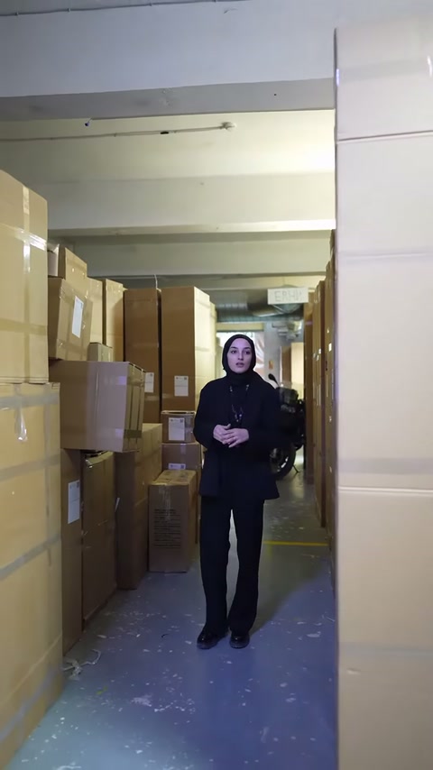 Depo koridorunda sunucu, istiflenmiş ürün kolileri arasında konuşuyor.