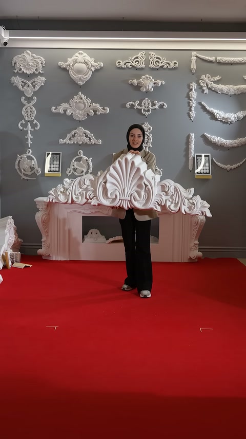 Presenter holds a large white decorative piece in front of ornamental wall panels.
