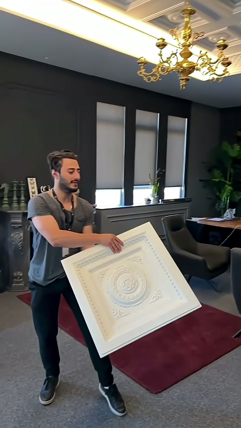 Presenter holds decorative ceiling panel with both hands toward viewer.