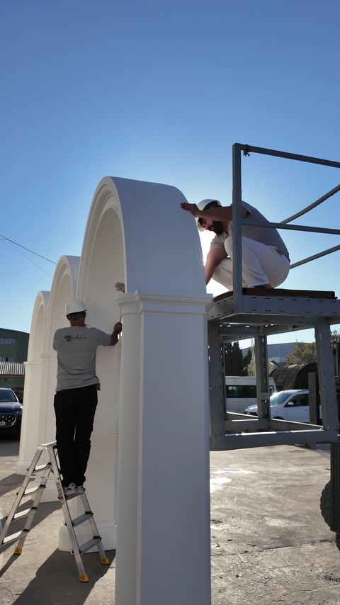 Workers install a large exterior arch using lifting equipment.