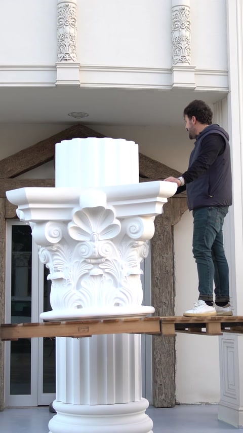 Un operaio su una piattaforma elevata posiziona un capitello sopra un fusto di colonna.