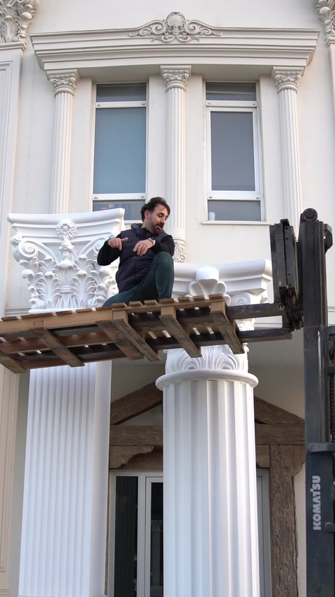 Una piattaforma di carrello elevatore solleva un operaio vicino a una facciata con colonne parzialmente installate.