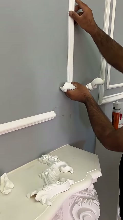 A curved white decorative corner element is being positioned against the corner of white trim on a gray wall