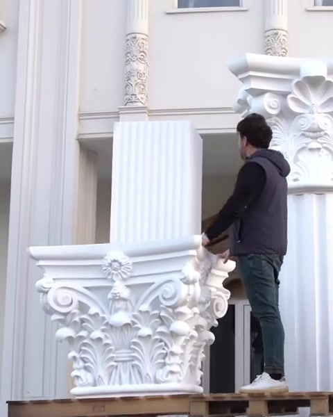 Worker holds Corinthian capital piece on wooden platform.