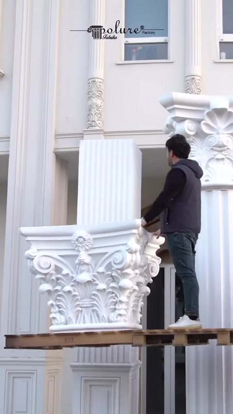 Worker holds Corinthian capital piece on wooden platform.