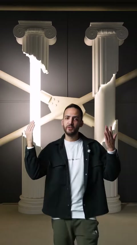 Presenter raises his hands before two illuminated classical columns in showroom.