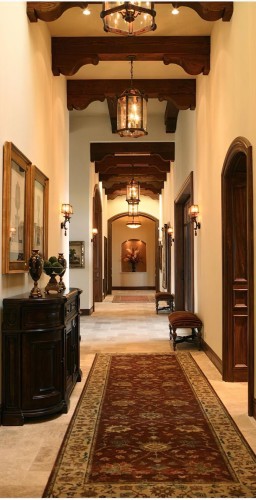 Rustic Classic Corridor with Ornamental Wood Details and Elegant Lighting <h2>Rustic Wooden Elements</h2><p>The corridor design emphasizes the beauty of <strong>wooden beams</strong> with sculpted curves and layered support details that create a rhythm along the ceiling. These elements are not only structural but decorative, reflecting the balance of functionality and artistry in classical architectural traditions.</p><h2>Lighting and Atmosphere</h2><p>Lantern-style <strong>pendant lighting</strong> hangs across the passage, casting a warm glow that enhances the golden hues of the wood and ivory-toned walls. Traditional <strong>wall sconces</strong> add accent lighting that highlights the framed artworks and architectural niches, contributing to a layered and intimate ambiance.</p><h2>Textile and Pattern Details</h2><p>A richly patterned <strong>runner carpet</strong> stretches the length of the corridor, balancing the verticality of the walls and beams with ornate floral and geometric motifs. Its earthy red background with beige and muted green detailing reflects classical rug traditions, adding visual warmth and grounding the space.</p><h2>Furniture and Decorative Touches</h2><p>The corridor features classic dark wood furniture, including carved consoles and benches with upholstered seats. A decorative vase arrangement at the end adds focal interest and connects with the organic motif of the carpet.</p><h2>Historical Note</h2><p>The use of wooden beams and carved details recalls medieval and Renaissance interiors, where ornamented ceilings were designed to showcase craftsmanship as much as structural efficiency. This design pays homage to that history while adapting it to modern interiors.</p>

<p>This rustic classic corridor design highlights detailed <strong>wooden beams</strong> with sculptural curves that create rhythm and depth. Lantern-style <strong>pendant lighting</strong> illuminates the hallway with a warm glow, complementing the rich wood and cream-toned walls. Traditional <strong>wall sconces</strong> accent the vertical surfaces, giving balance and intimacy. A detailed <strong>runner carpet</strong> stretches elegantly, decorated with floral and geometric motifs in earthy hues. The furniture includes ornate consoles and benches that emphasize craftsmanship. The arrangement of framed artworks reinforces the corridor’s symmetry. The use of rich wood tones contrasts beautifully with the pale stone floor. Arched doorways create architectural fluidity throughout the corridor. Historical influence is visible in the woodwork’s decorative carvings. The interplay of dark furniture and soft light creates harmony. Every surface contributes to a sense of timeless elegance. Decorative niches with vases add focal points. The patterned carpet softens the stone while guiding movement. Ceiling beams emphasize traditional craftsmanship. The overall corridor atmosphere merges rustic tradition with refined classical design.</p>