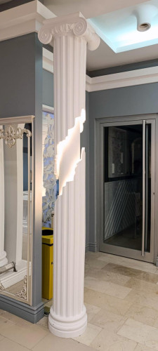 Polyurethane Decorative Illuminated Column for Neoclassical Living Room Design <h2>Neoclassical Elegance with Polyurethane Columns</h2><p>The use of <strong>polyurethane</strong> decorative columns has become increasingly popular in interiors designed to convey both durability and refined aesthetic qualities. This specific design, featuring an Ionic-style capital with coiled volutes, brings a sense of timeless grace into a salon setting.</p><p>The column showcases deep vertical <strong>fluting</strong> along its shaft, an element historically associated with the grandeur of classical temples. A modern twist is achieved through a sharp-edged illuminated opening, casting a gentle glow that highlights surrounding surfaces while serving as a functional artistic detail.</p><p>In neoclassical interiors, symmetry, ornamentation, and balance are fundamental. The integration of a glowing accent within the column delivers contrast, blending <strong>traditional</strong> detail with contemporary innovation. The geometric cut disrupts uniformity in a deliberate design choice, allowing light to transform the column into both a structural reference and a visual centerpiece.</p><h2>Design Details and Inspiration</h2><p>The polished white finish emphasizes harmony with surrounding gray wall panels and mirrored frames. Reflective surfaces enhance the lighting effect, ensuring that the column does not merely support but also defines the overall spatial narrative. The motifs align with <strong>classical</strong> decoration yet reveal a distinctly modern sensibility optimal for personalized interiors.</p><p>Historically, Ionic and Corinthian columns of ancient Greece applied symmetry and proportion as essential design language. By reinterpreting these motifs in a polymer medium, this piece achieves lighter weight, durability, and customization potential, making it an inventive fit for a living room or reception space.</p><ul><li>Polyurethane construction ensures durability and easy installation.</li><li>Illuminated cut pattern provides both function and artistic depth.</li><li>Neoclassical style enriches a salon design with elegance and grandeur.</li></ul><p>Through the play of light, geometry, and ornament, the design blends <strong>neoclassical</strong> sophistication with a distinctly modern identity.</p>
<p>This polyurethane column combines classical design with modern illumination. The Ionic-inspired capital provides timeless elegance. Vertical fluting emphasizes height and refinement. A geometric cut with hidden light transforms the structure into a glowing feature. The design suits salons, living rooms, and reception areas. White surfaces contrast with gray walls for a clean aesthetic. Reflective mirrors amplify the soft glow. The illuminated feature enhances atmosphere without excess. Classical motifs link the piece to architectural history. Polyurethane ensures strength and practicality. Decorative grooves balance traditional and modern harmony. The illuminated cut adds contemporary depth. This column is both structural and sculptural. It celebrates <strong>neoclassical</strong> inspiration while offering <strong>modern</strong> function. A <strong>decorative</strong> column becomes a centerpiece of design.</p>